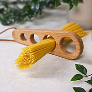 Spaghettimaß aus Eichenholz mit Lederband und eingesteckten Spaghetti, dazu Blumen im Hintergrund