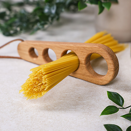 Spaghettimaß aus Eichenholz mit Lederband und eingesteckten Spaghetti, dazu Blumen im Hintergrund